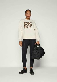 Krém színű pulóver leopárdmintás "DKNY" felirattal, fekete leggingszel és fekete sportcipővel párosítva. Fekete tornazsákot tart a kezében.