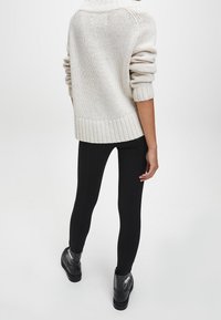 Kremowy sweter dzianinowy z podwiniętymi mankietami, ściągaczem na dole, zestawiony z czarnymi dopasowanymi leginsami i czarnymi botkami za kostkę. Zwróć uwagę na teksturę i warstwowość.