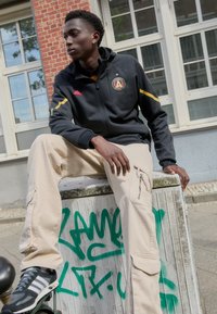 Schwarze Zip-Jacke mit gelben Streifen und Logo, kombiniert mit beigen Cargo-Hosen mit Reißverschlusstaschen. Graue Sneakers mit weißen Streifen.