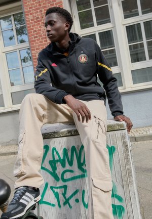 Zwarte zip-up jas met gele strepen en logo, gecombineerd met beige cargobroek met ritssluitingen. Grijze sneakers met witte strepen.