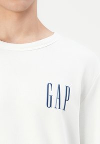 Sudadera blanca con cuello redondo que presenta un logo "GAP" en azul marino en el pecho. La tela parece suave y ligeramente texturizada.