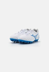 Botas de fútbol blancas con tacos, cordones y acentos azules, posicionadas una al lado de la otra sobre un fondo blanco.