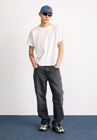 White, short-sleeve t-shirt; dark gray jeans; blue denim cap; black sunglasses; patterned sneakers; tattoos visible on arms.