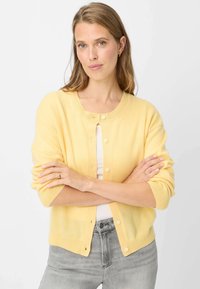 Gelber Cardigan aus weichem Material mit rundem Ausschnitt, Knopfverschluss und dreiviertel Ärmeln, kombiniert mit grauen Jeans.
