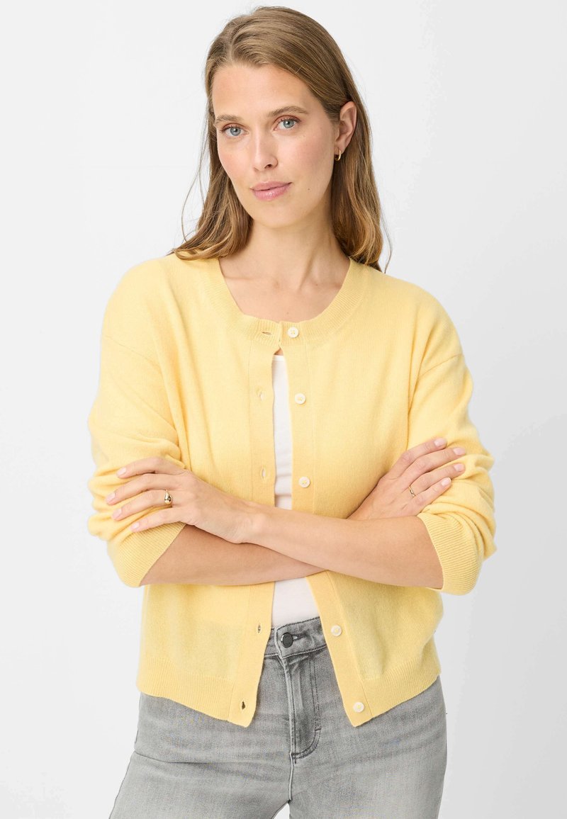 Gelber Cardigan aus weichem Material mit rundem Ausschnitt, Knopfverschluss und dreiviertel Ärmeln, kombiniert mit grauen Jeans.
