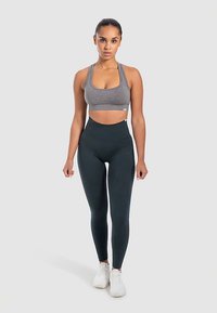 Sujetador deportivo gris y leggings oscuros de talle alto. Textura suave, diseño ajustado y costuras reforzadas. Modelo de pie con zapatillas blancas.