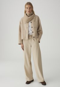 Frau trägt beige geknöpfte Jacke mit großen Taschen, passende weit geschnittene Hose, einen Schal, weißes Oberteil und braune Schuhe, steht vor einem einfarbigen Hintergrund.