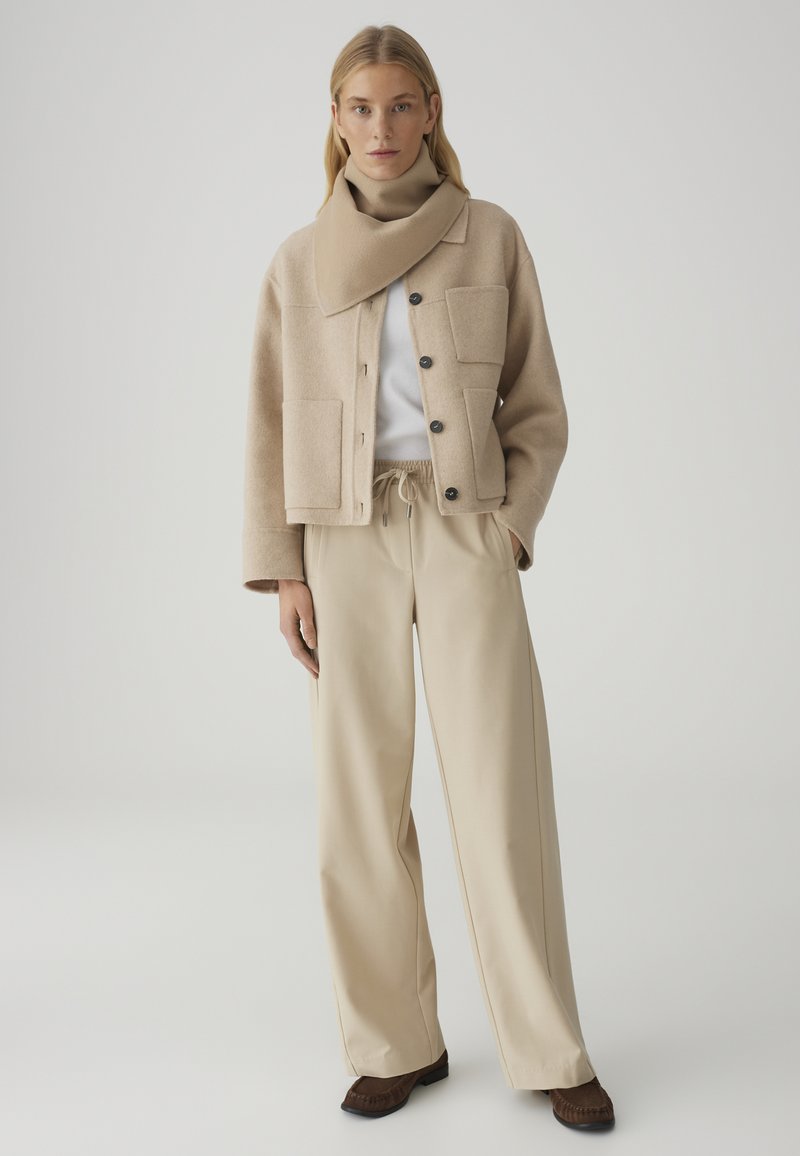 Frau trägt beige geknöpfte Jacke mit großen Taschen, passende weit geschnittene Hose, einen Schal, weißes Oberteil und braune Schuhe, steht vor einem einfarbigen Hintergrund.