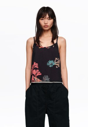 Młoda kobieta z prostymi czarnymi włosami nosi czarny bezrękawnik z kolorowymi wzorami koralowymi oraz czarne spodnie, mając ręce w kieszeniach.