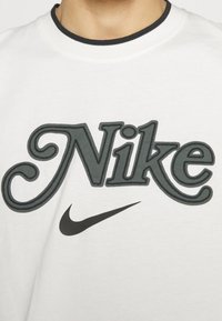 Vit bomull t-shirt med en stor mörkgrå "Nike" logotyp med dekorativ sömnad och svart Swoosh under. Rund halsringning med svart kant.