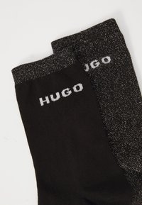 Black socks with a textured, glittery cuff and knitted body. Features "HUGO" logo in white on each sock. Pair of contrasting designs.