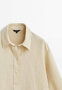 Chemise beige en lin à manches longues, avec col et boutonnage devant, présentée sur un fond blanc.