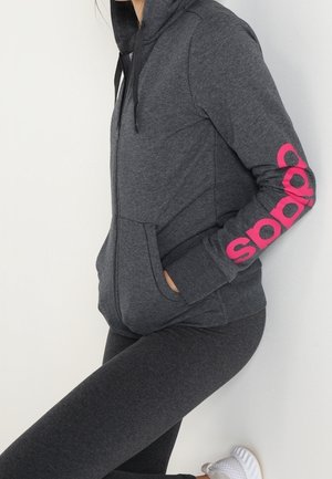 Osoba nosząca ciemnoszarą bluzę z kapturem i zamkiem błyskawicznym z różowym napisem „adidas” na rękawie, dopasowane legginsy i białe buty sportowe, opierająca się o ścianę.