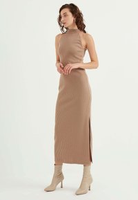 Ribbad, ärmlös midi-klänning i tan med hög halsringning och sidoklyfta, parat med beige ankelstövlar med spetsig tå.