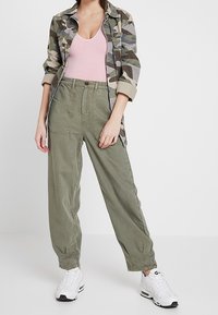 Camo-patterned shirt jacket, light pink tank top, olive green baggy pants, and white sneakers. Relaxed fit with tapered cuff details.