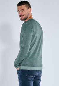 Grüner, strukturierter Pullover mit langen Ärmeln und rundem Halsausschnitt, hinten mit einem Knopf versehen. Kombiniert mit dunklen Jeans.
