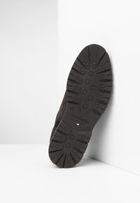 Scarpa in suede marrone con suola in gomma texturizzata, caratterizzata da un battistrada decorato e un marchio sulla suola, inclinata contro uno sfondo bianco.