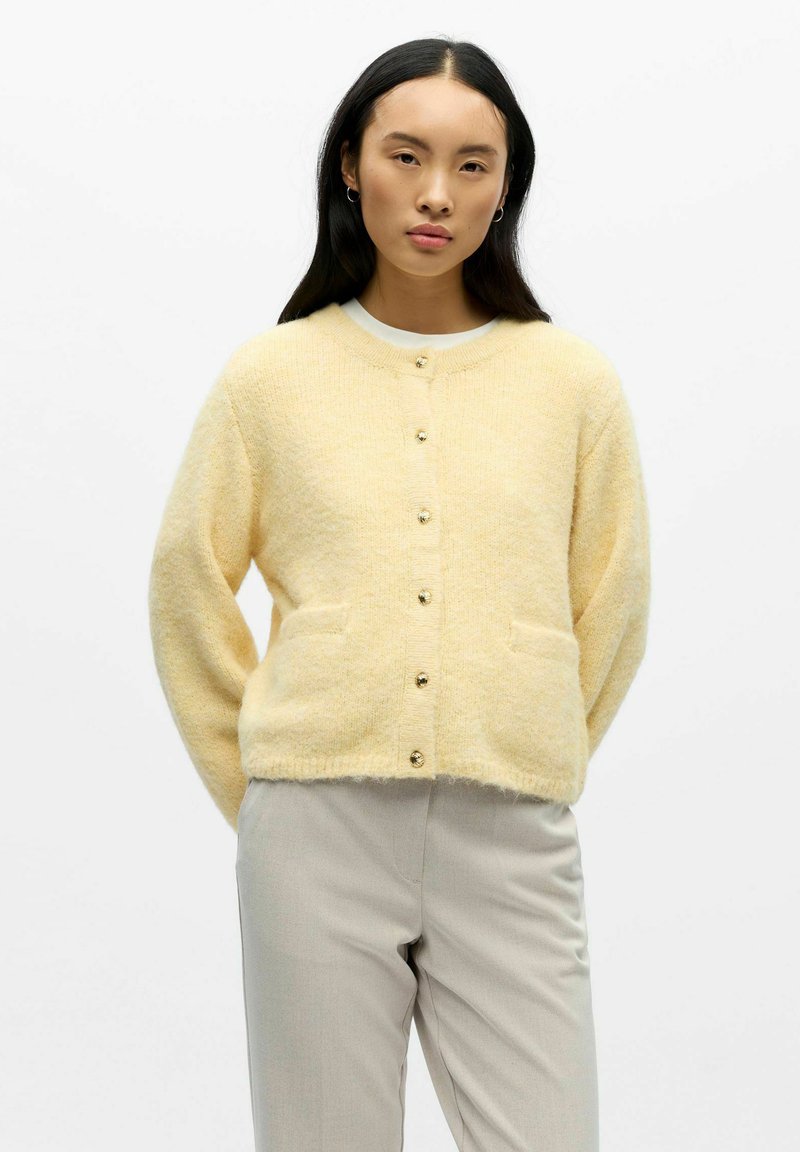 Cárdigan amarillo de textura suave con escote redondo, botones delanteros y dos bolsillos, combinado con pantalones de color claro. Detalle del tejido texturizado visible.