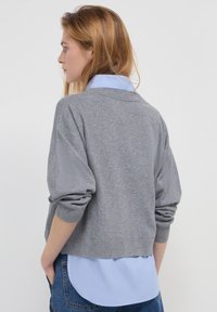 Maglione grigio lavorato a maglia con scollo rotondo e taglio cropped, indossato sopra una camicia colletto azzurro chiaro, con maniche arrotolate e una consistenza liscia.