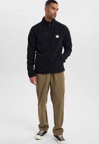 Giacca in pile nera con zip completa, colletto alto e patch con logo. Indossata sopra pantaloni cargo beige e sneakers nere. Tessuto liscio e caldo.