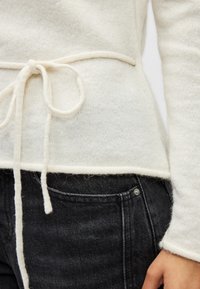 Gros plan sur une taille de pull en maille crème noué, portée sur un jean en denim noir avec une poche avant visible et une main reposant à côté.