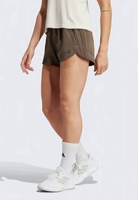 Καφέ αθλητικά σορτς με χαλαρή εφαρμογή, με πλαϊνές σχισμές και διακριτές κατακόρυφες ρίγες, συνδυασμένα με λευκά αθλητικά παπούτσια και κάλτσες.