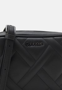 Czarna pikowana skórzana torba z regulowanym paskiem, zapinana na zamek, z tłoczonym logo Calvin Klein na przodzie. Gładka tekstura.