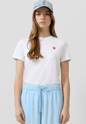 Vrouw met een lichtblauwe baseballpet, wit T-shirt met een klein rood aardbeiembleem, en lichtblauwe broek met trekkoord, staand tegen een effen achtergrond.