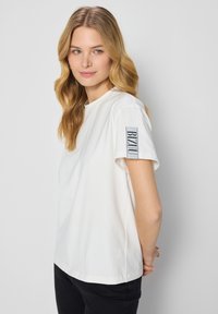 Biała koszulka krótkiego rękawa z okrągłym dekoltem i czarnym logo "BIZOU" na lewym rękawie. Miękka faktura, luźny krój, zestawiona z czarnymi spodniami.