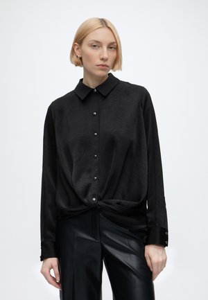 Blonde Frau trägt eine schwarze Bluse mit Knöpfen und Knoten an der Taille sowie schwarze Lederhose, steht vor einem schlichten weißen Hintergrund.