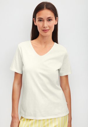 Woman with long brown hair wearing a plain white V-neck T-shirt and yellow striped skirt against a light gray background.