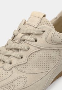 Μπεζ αθλητικό παπούτσι από σουέντ με διάτρητες λεπτομέρειες, επίπεδα κορδόνια, ενισχυμένη μύτη και λαστιχένια σόλα. Κοντινή όψη που εστιάζει στην υφή και το σχέδιο.