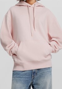 Felpa con cappuccio in pile rosa chiaro con tasca frontale a marsupio, cappuccio con coulisse e polsini e orlo a coste, abbinata a jeans azzurri.