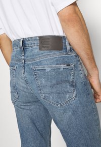 Homme portant un jean en denim bleu clair avec une étiquette de marque en cuir marron et une chemise blanche à manches courtes, vu de dos, de la taille vers le bas.