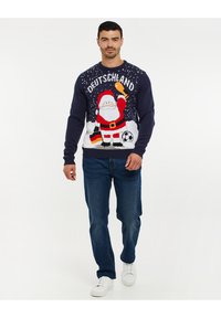 Gebreide marineblauwe trui met een feestelijk kerstmanontwerp, voorzien van de tekst "DEUTSCHLAND", sneeuwvlokken en een patroon van een voetbal; gecombineerd met blauwe jeans.