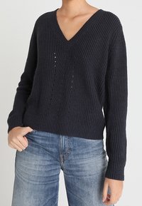 Pull en tricot bleu marine avec un col en V, des manches longues, une texture ribbed et des détails découpés subtils à l'avant, assorti à un jean en denim clair.