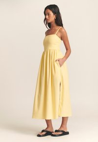 Keltainen, hihaton maksimimekko, jossa on vartalonmyötäinen yläosa, laskeutuva helma, sivutaskut ja halkioyksityiskohta, valmistettu pehmeästä kankaasta.