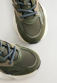 Žalios sportinės bateliai, pagaminti iš tinklelio ir zomšos medžiagų, su kontrastingomis pilkomis raištelėmis ir storu baltu padu. Tekstūruoti ir išlenkti dizaino elementai.
