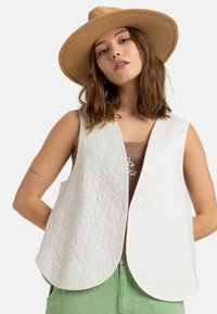 Chaleco acolchado blanco con un patrón floral, de frente abierto y bordes redondeados, llevado sobre una camiseta marrón con pantalones de cintura alta verdes.