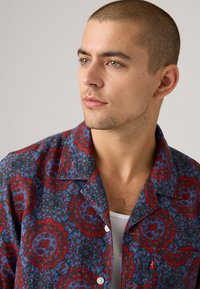 Man with short hair wearing a red and blue ornate patterned button-up shirt over a white ribbed tank top, looking right.