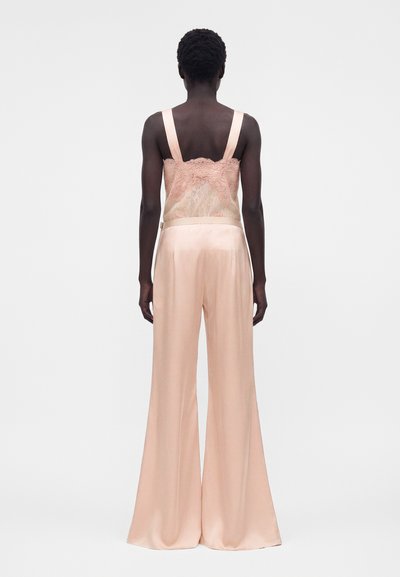 Γυναίκα με κοντά σγουρά μαλλιά, ντυμένη με ανοιχτό ροζ δαντελένιο καμησόλ και φαρδιά σατέν παντελόνια, εμφανιζόμενη από πίσω σε μονόχρωμο φόντο.