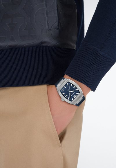Tengerészkék pulóver bordázott mandzsettákkal, texturált bőrpanellel díszítve. A csuklón egy nagy ezüst-kék óra, amely a modern dizájnt és a funkcionalitást ötvözi.