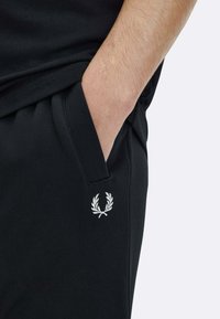 Schwarze Sporthose mit einer Seitentasche und einem kleinen weißen, bestickten Lorbeerlogo auf dem linken Oberschenkel. Glatte Textur, elastischer Bund.