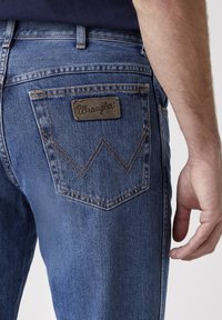 Vaqueros de mezclilla en azul medio con textura desgastada, con una etiqueta de cuero de Wrangler y bolsillos traseros doblemente cosidos con un patrón en zigzag.