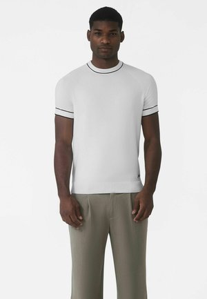 Bílý pletený svetr s krátkými rukávy a černým lemováním u krku a rukávů, s přiléhavým střihem a hladkou texturou. Nošený s lehkými kalhotami.