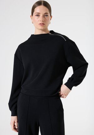 Sweat-shirt noir à col montant, à manches longues et avec un détail de fermeture éclair sur le côté. Texture lisse et coupe décontractée.