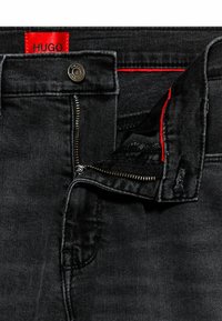 Vaqueros de mezclilla negros con cremallera parcialmente desabrochada, botón de metal y etiqueta roja de la marca HUGO en la cintura interna.