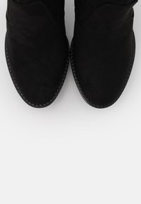 Bottines en daim noir avec un bout arrondi, des accents cousus le long de la semelle et une texture douce. Elles présentent un design minimaliste et classique.