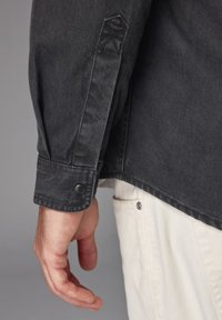 Camicia in denim nero con colletto a punta, polsini abbottonati e cuciture diagonali sulle spalle, abbinata a pantaloni chiari e una tasca laterale.