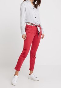 Jean en coton rouge avec une coupe ajustée, dotée d'une ceinture à nouer et de coutures contrastantes, portée avec une chemise à rayures à manches longues et des baskets blanches.